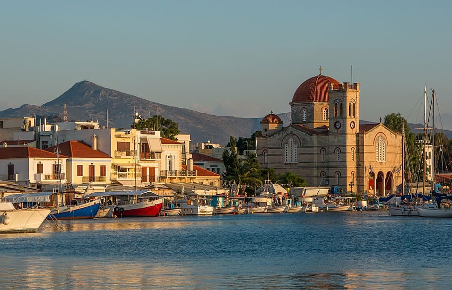 A photograph of the port at the Greek island of Aegina in the evening sun. The foreground shows blue sea with some medium and small-sized boats. Behind, there are some houses with balconies and awnings, a church with a tower and a dark brown dome. In the distance, there is a mountain. 